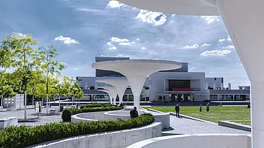 Die Aufnahme zeigt den Vorplatz des Staatstheaters Darmstadt. Moderne Architektur mit Grünflächen und Fußgängern.