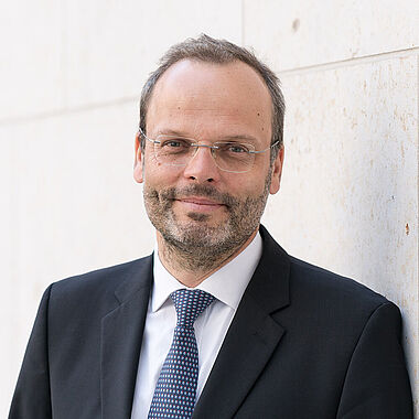 Professioneller Mann in Anzug und Krawatte, lächelnd vor neutralem Hintergrund.