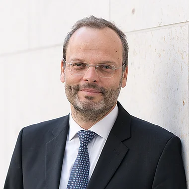 Professioneller Mann in Anzug und Krawatte, lächelnd vor neutralem Hintergrund.