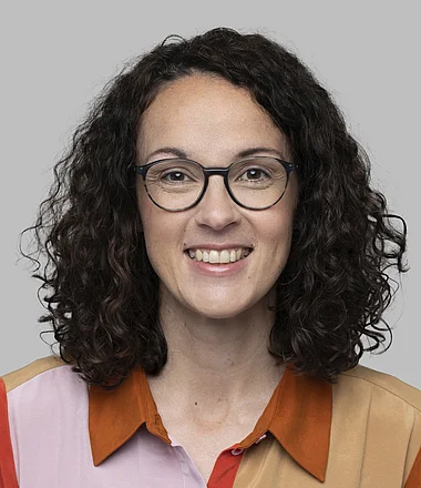 Eine lächelnde Frau mit lockigem Haar und Brille trägt ein farbiges Hemd.