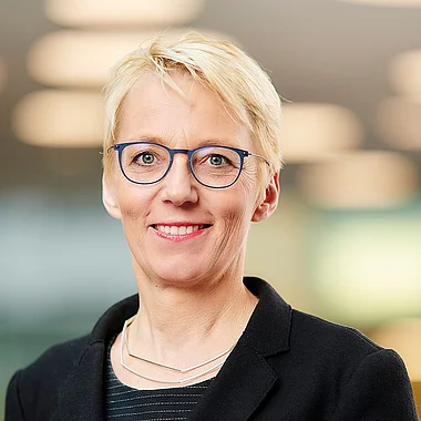 Frau in Anzug, lächelnd, vor moderner Büroarchitektur.