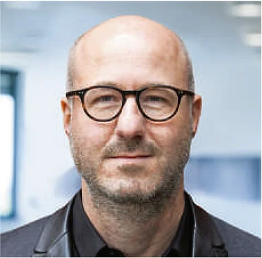 Ein Mann mit Bart und Brille trägt einen dunklen Anzug.